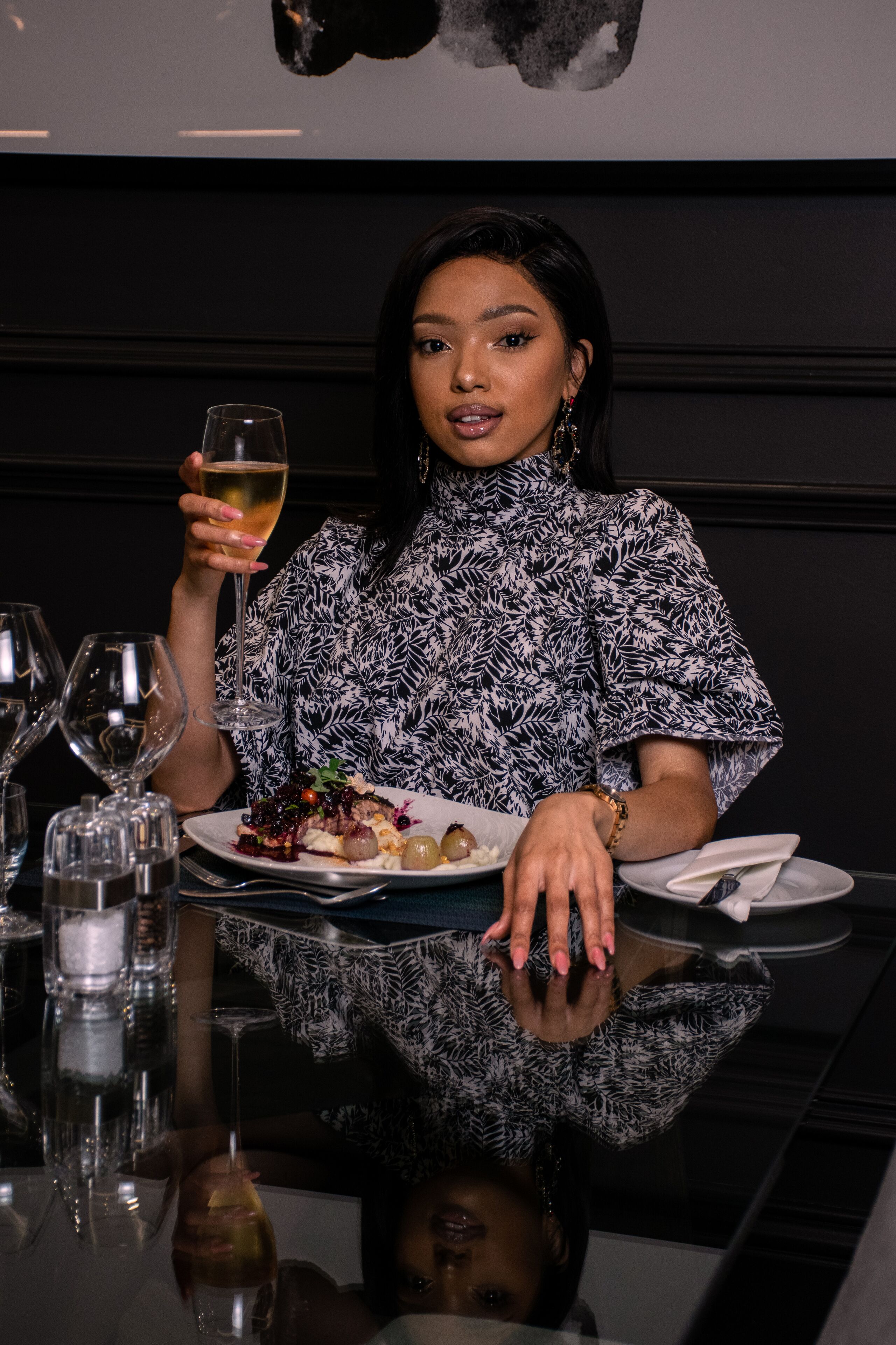 A person seated at a table with a glass of wine, a plate of food, and a reflection on the table surface.