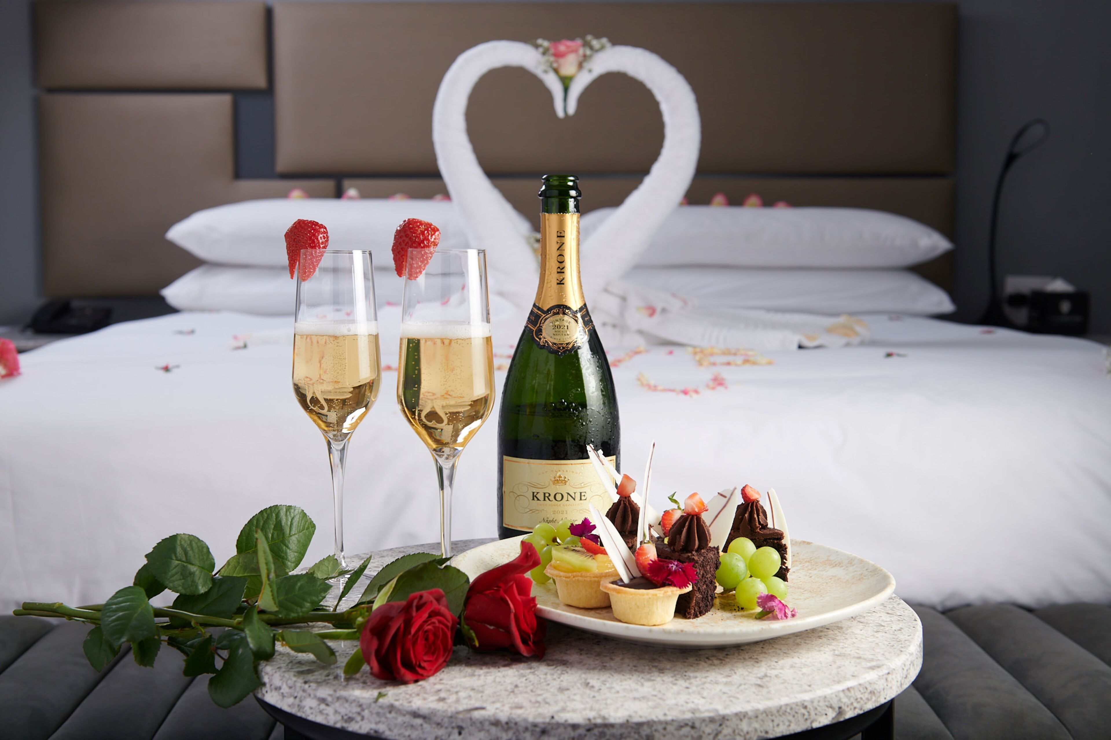 A bed with a heart-shaped decoration, a bottle of Krone champagne, two glasses of champagne, and a plate with various desserts.