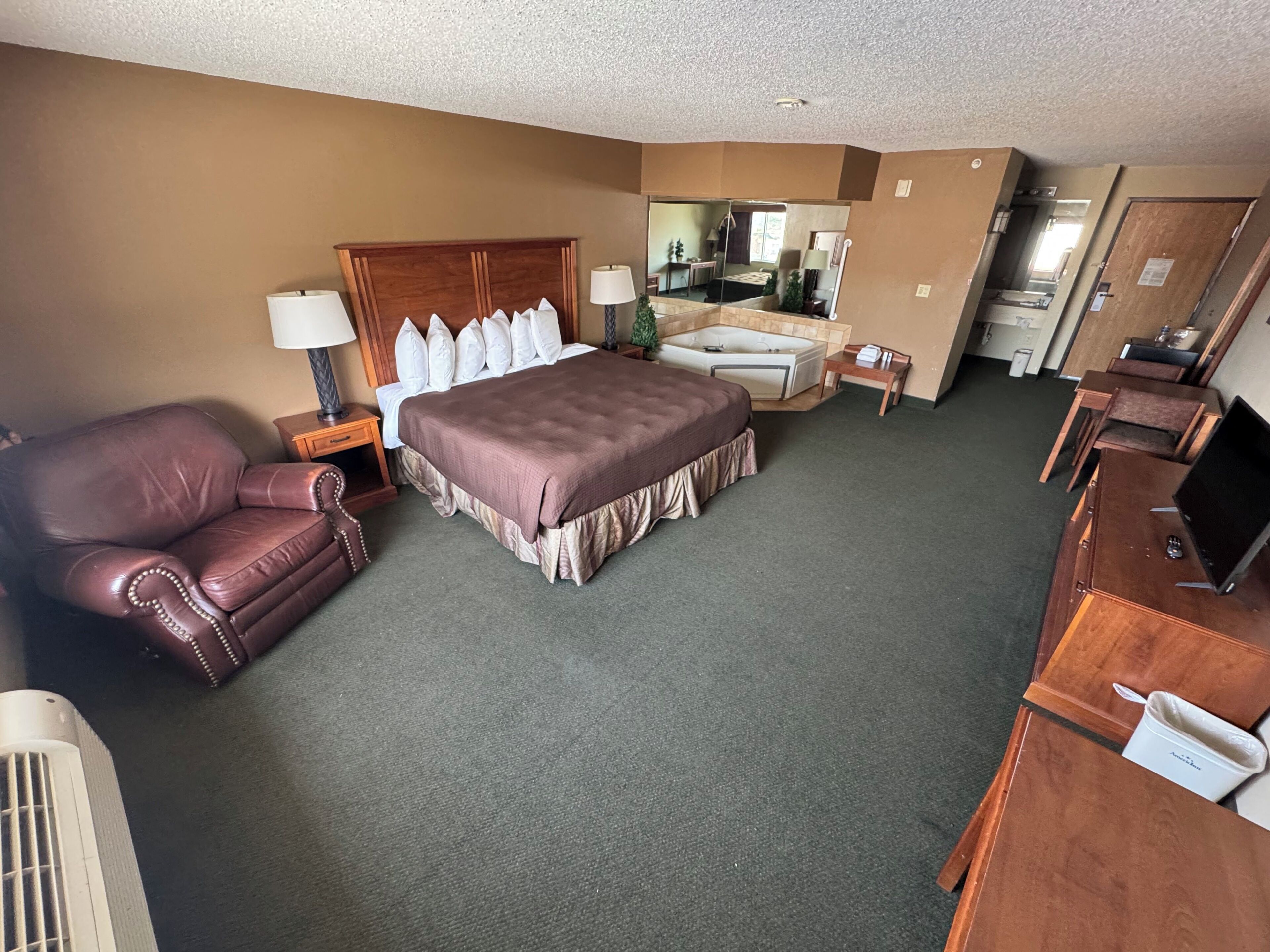 Une chambre d’hôtel avec un grand lit, un fauteuil en cuir, un bureau avec un ordinateur, une petite table avec une lampe et une salle de bain visible à travers une porte.