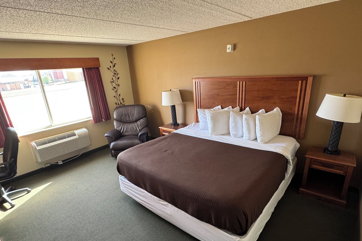 A hotel room with a large bed, two bedside tables with lamps, a chair, a window with curtains, and a wall-mounted air conditioner.