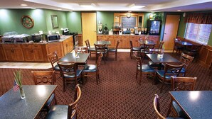 A restaurant with wooden tables and chairs, a kitchen area, and a patterned carpet.