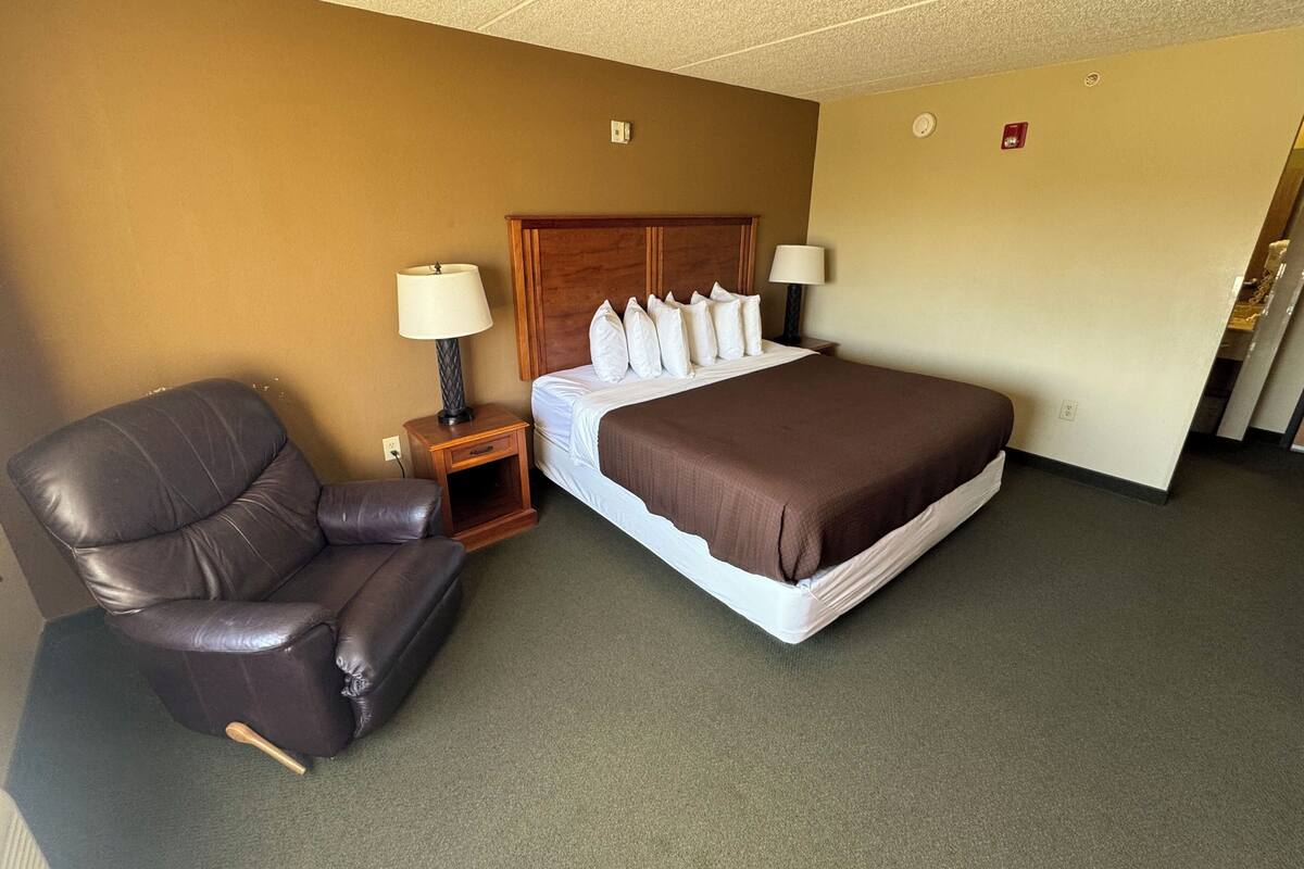 A hotel room with a bed, a leather chair, two bedside tables with lamps, and a carpeted floor.