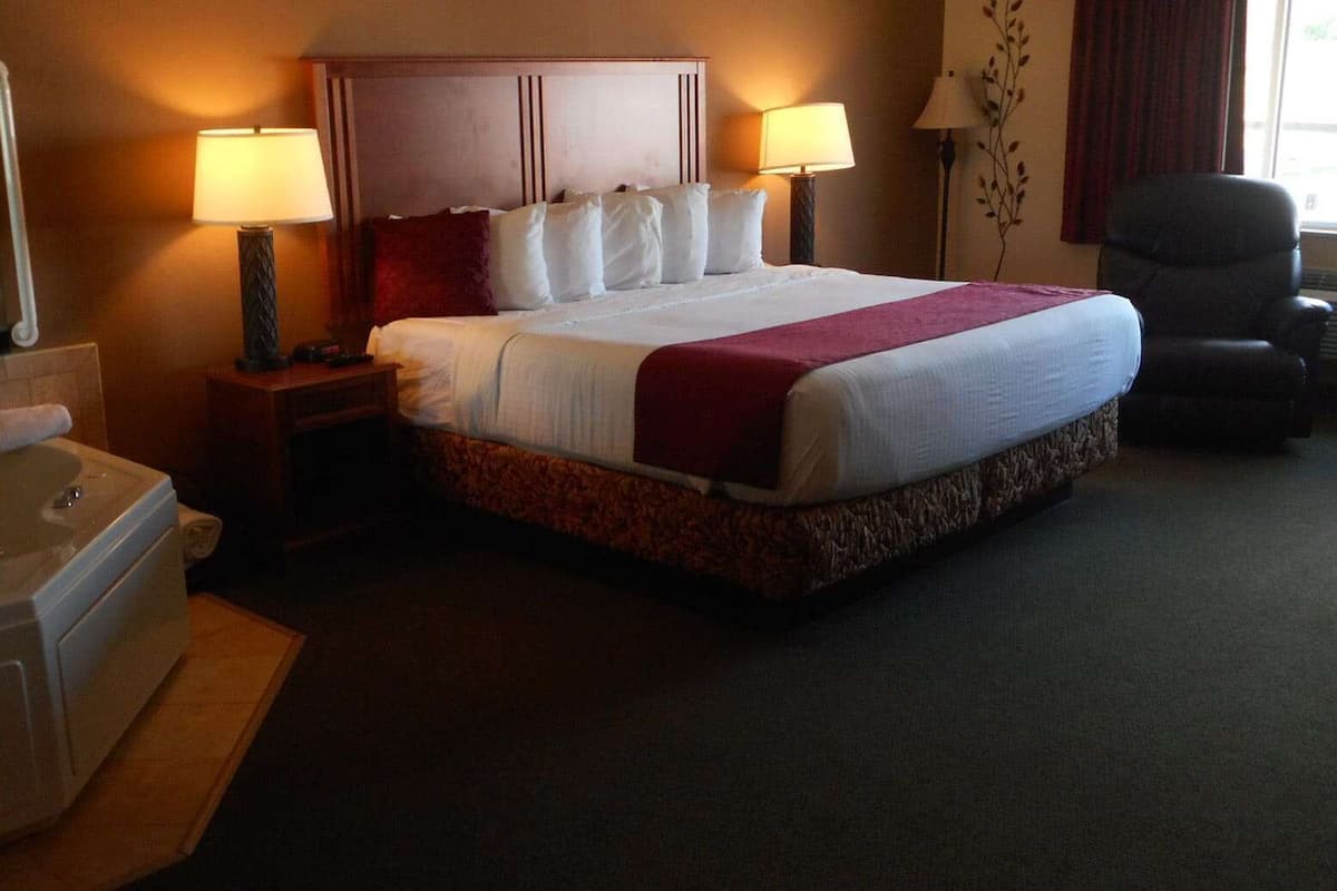 A hotel room with a large bed, two bedside lamps, a chair, a desk with a lamp, and a bathtub.
