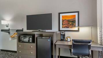 Quarto de hotel com televisão de tela plana, geladeira, micro-ondas, escrivaninha, cadeira, cafeteira e quadro decorativo na parede.