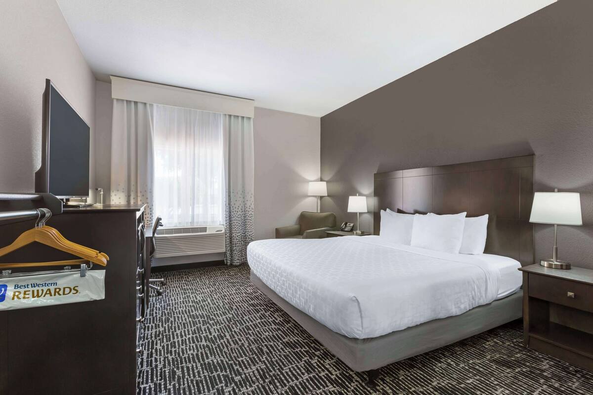 Ein Hotelzimmer mit einem großen Bett, zwei Nachttischen inklusive Lampen, einem Schreibtisch mit Stuhl, einem Kleiderständer und einem Fenster mit Vorhängen.