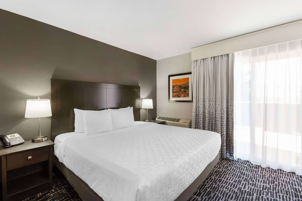 Ein Hotelzimmer mit einem großen Bett, zwei Nachttischlampen, einem Telefon, einem gerahmten Bild an der Wand und einem Fenster mit transparenten Vorhängen.