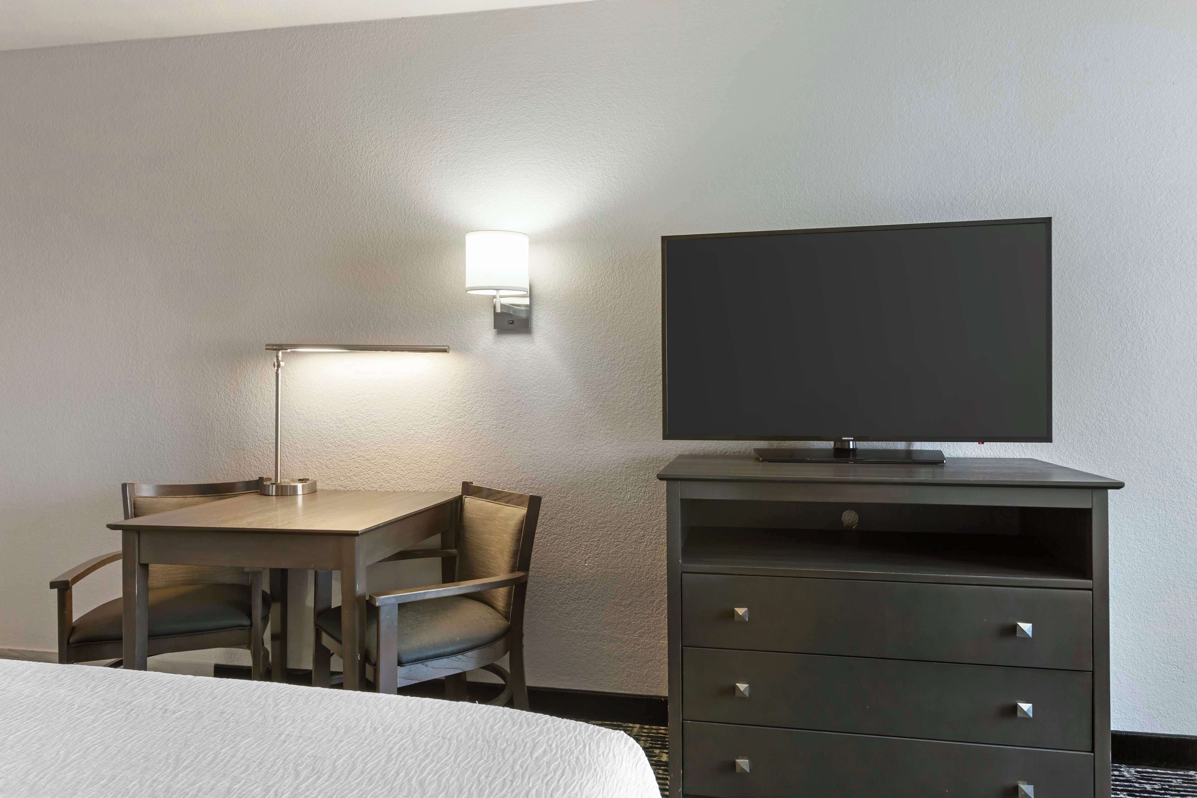 A hotel room with a bed, a desk with a lamp, a TV on a dresser, and a wall-mounted light.