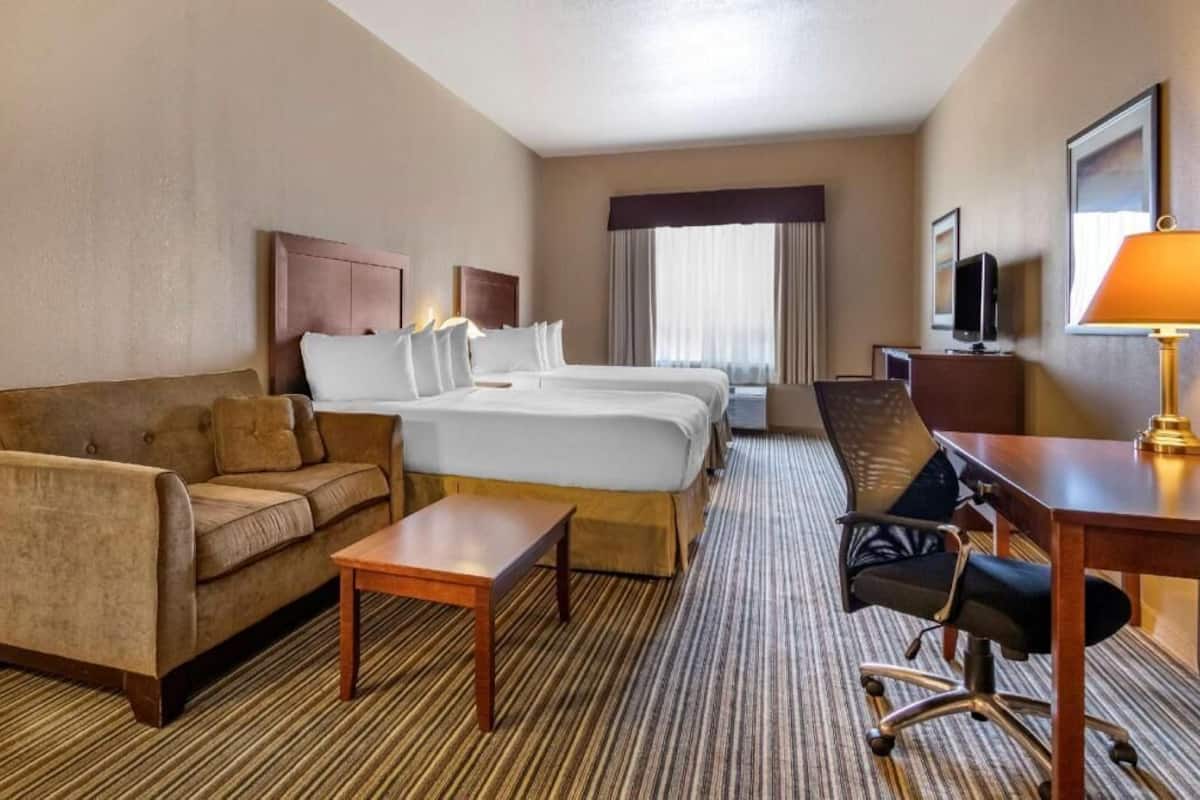 A hotel room with a sofa, two beds, a desk, a chair, a TV, and a lamp.