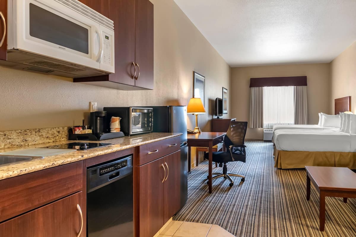 A hotel room with a kitchenette, including a microwave, coffee maker, and dishwasher. There is a bed, a desk with a chair, and a TV.