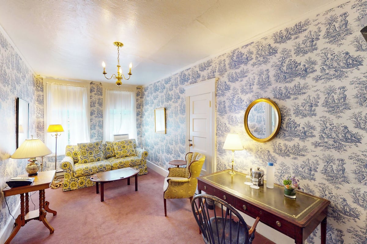 A room with a patterned wallpaper, a chandelier, a sofa, a chair, a table, and a mirror.