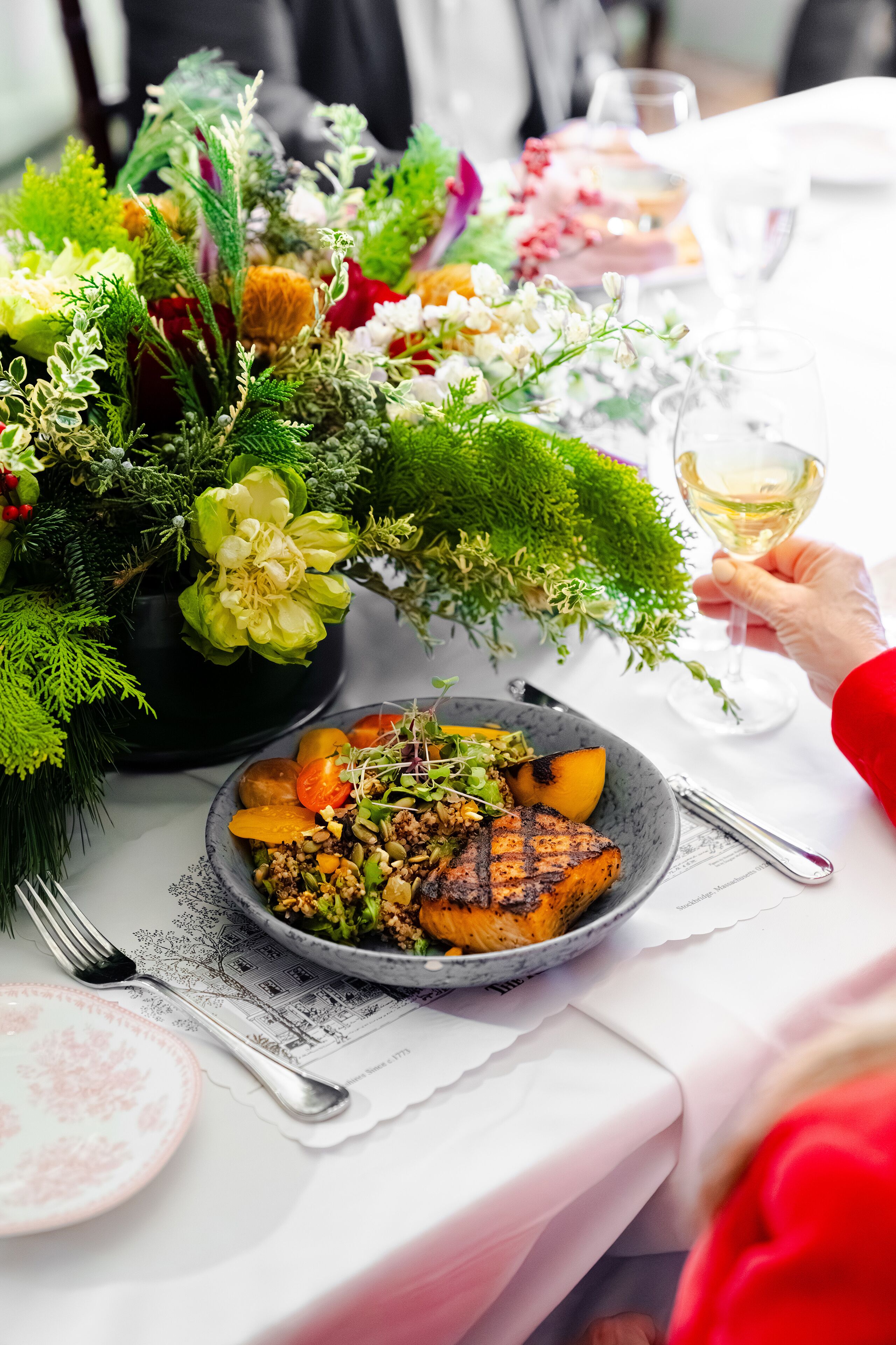 A dining table with a plate of food, a glass of white wine, and a decorative floral arrangement.