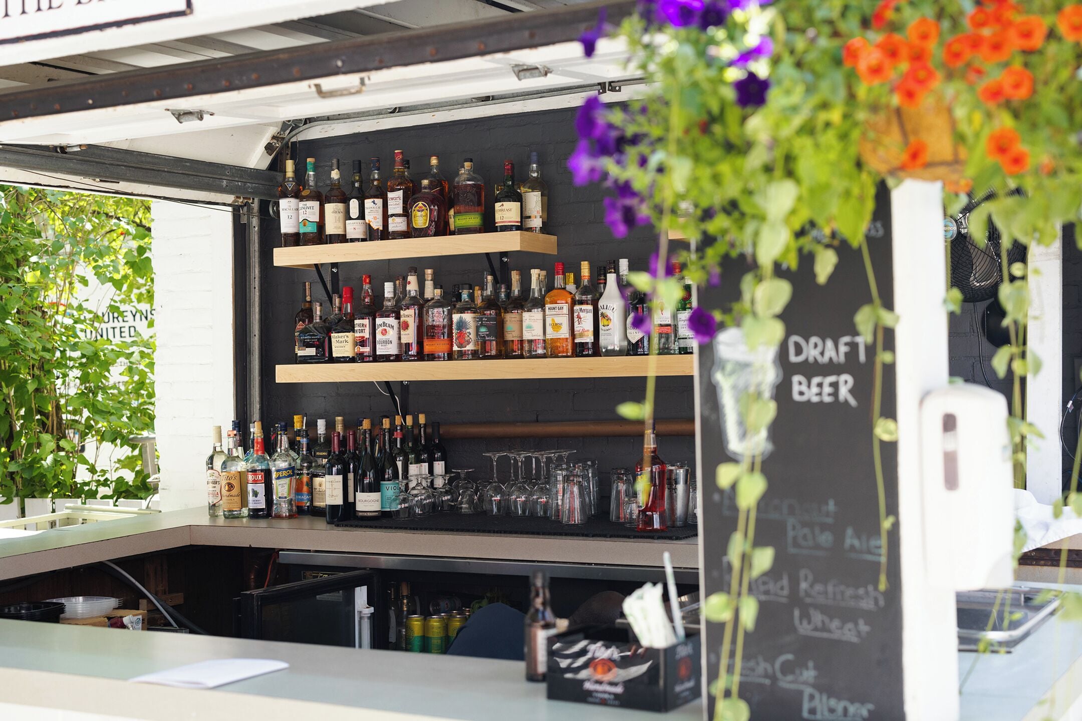 A bar area with shelves of liquor bottles and glasses, a chalkboard menu, and a hanging flower arrangement.