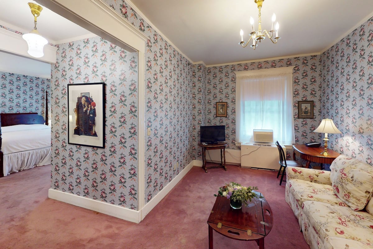 A room with floral wallpaper, a bed, a television, a desk with a laptop, a sofa, and a chandelier.