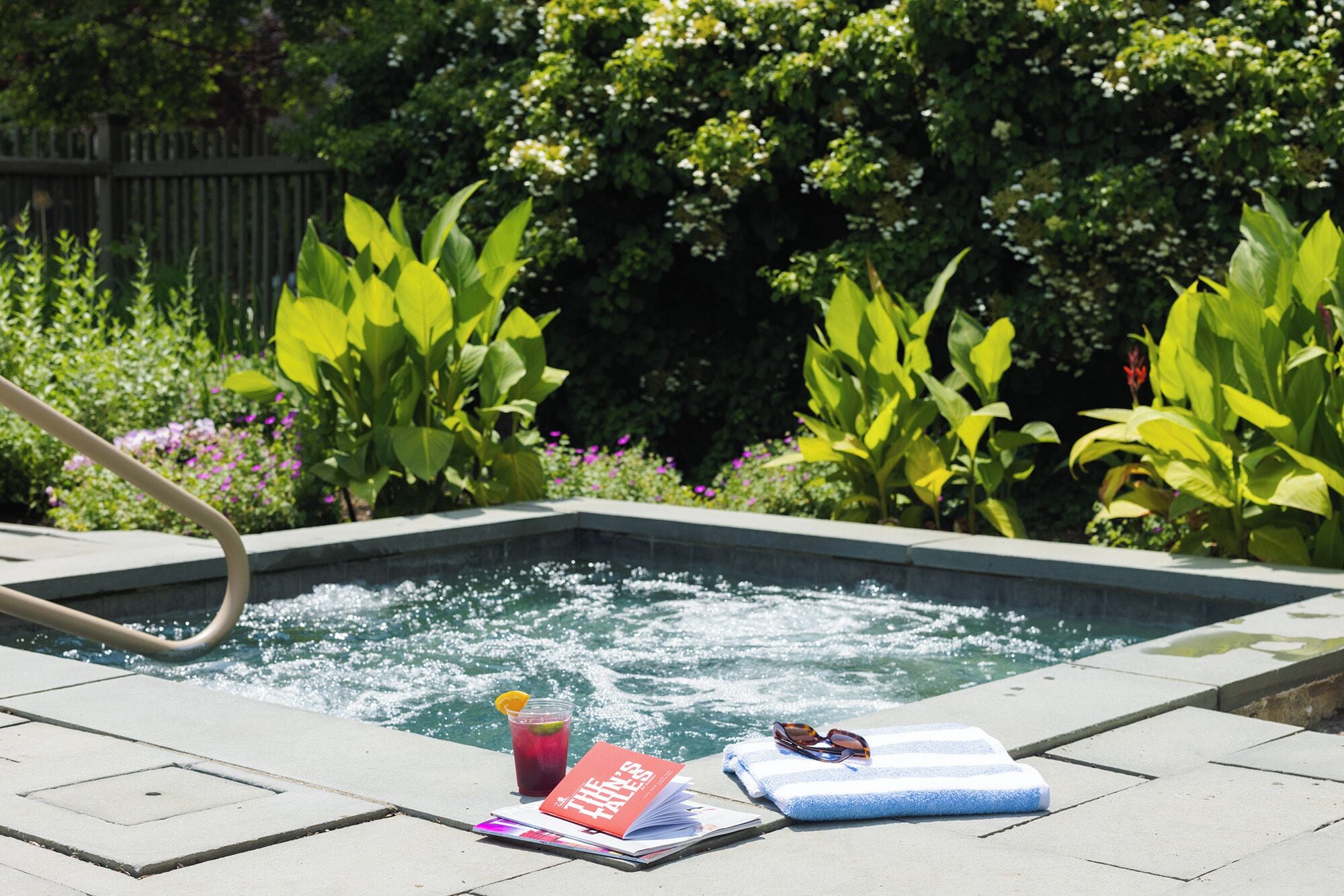 A swimming pool with a water fountain, surrounded by lush greenery and a wooden fence.