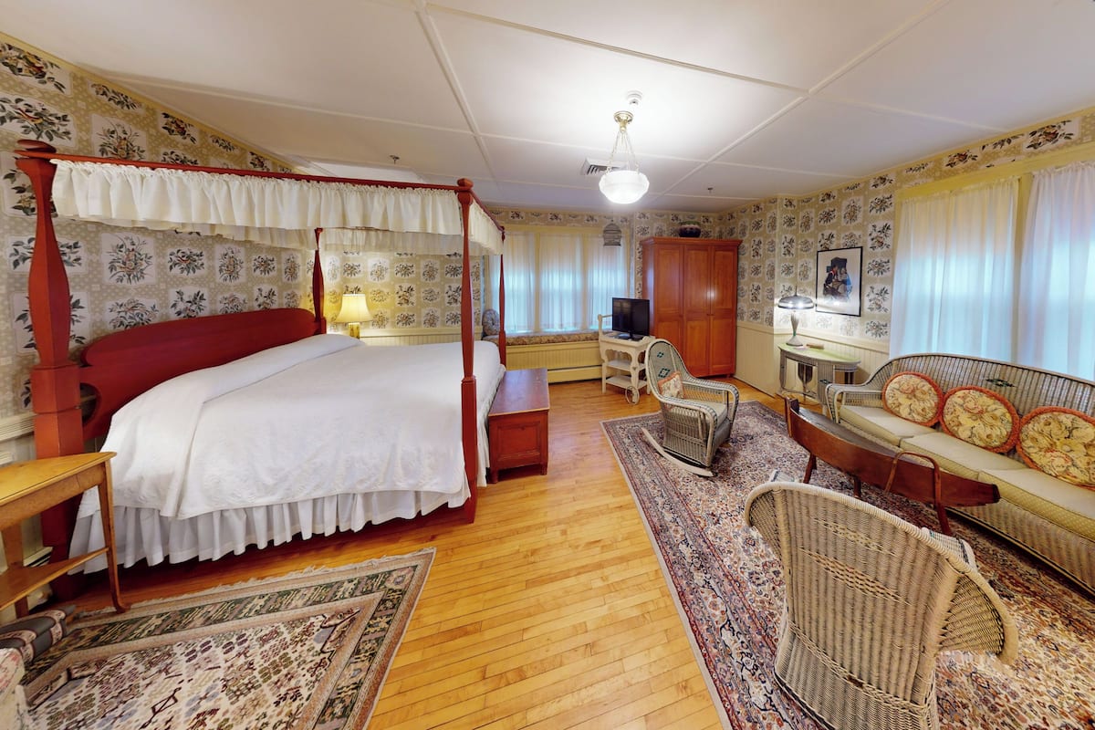 A traditional bedroom with a four-poster bed, a wooden desk, a wicker sofa, and a patterned rug.