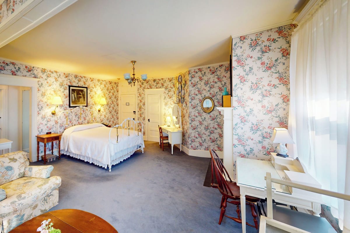 A bedroom with floral wallpaper, a bed, a desk, a chair, and a dresser.