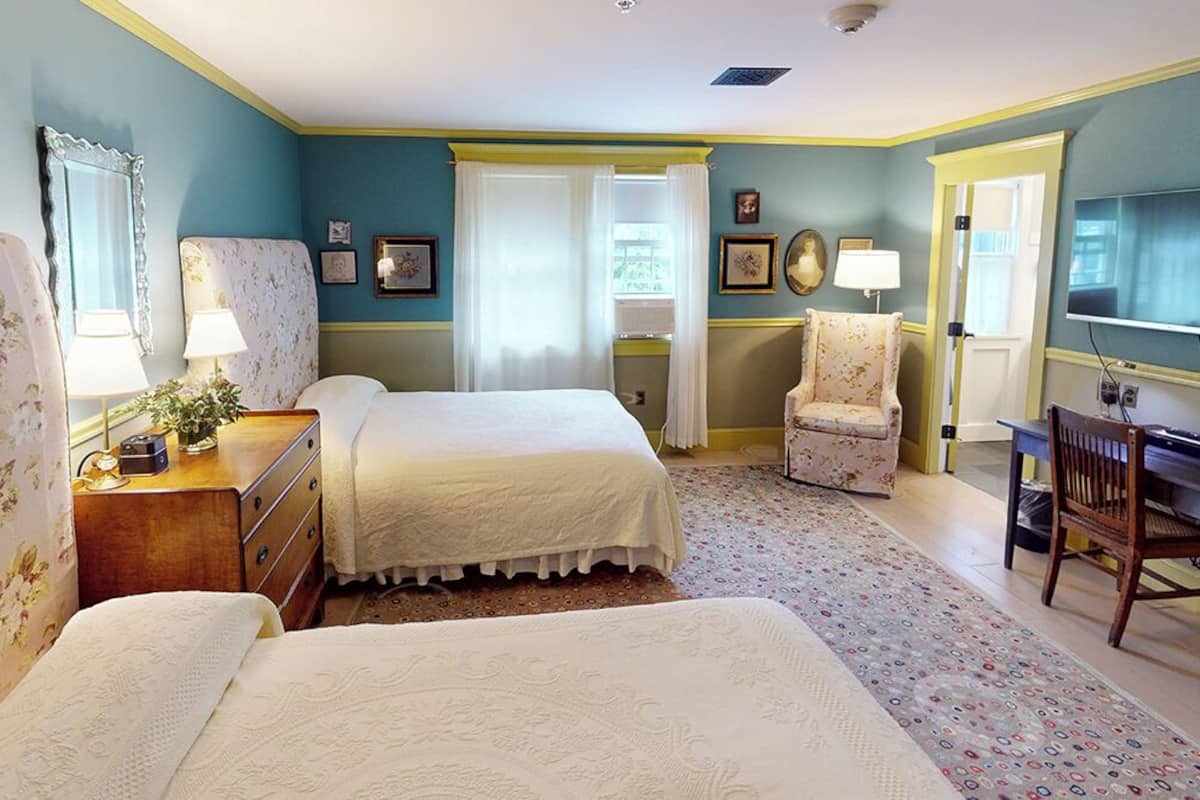 A bedroom with two beds, a desk, a chair, a television, and a window with curtains.
