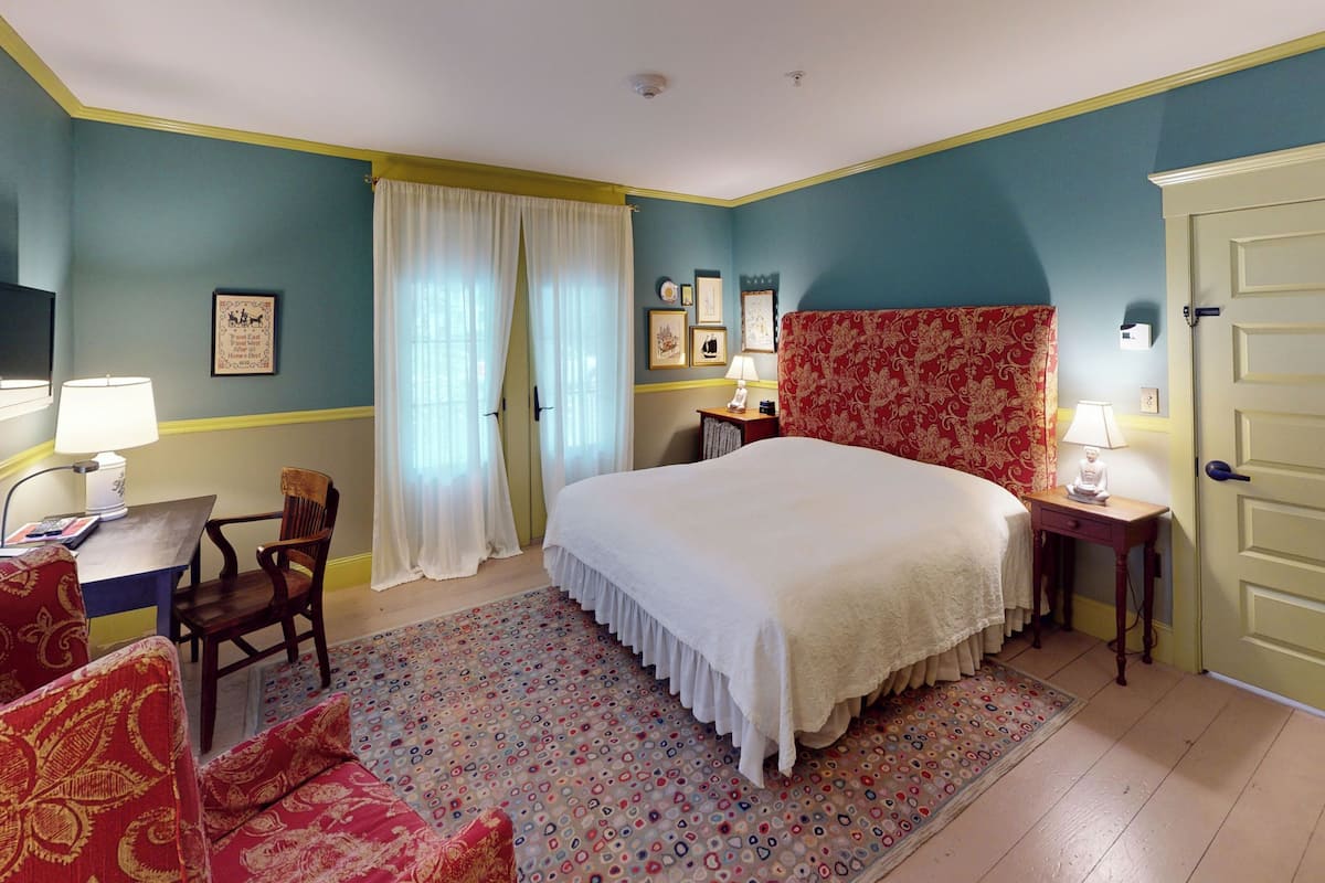 A bedroom with a large bed, a desk with a chair, a small table with a lamp, and a door.