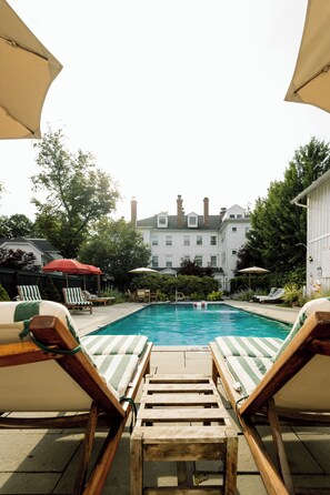 Un espace piscine avec des chaises longues et des parasols, entouré de verdure et avec un bâtiment en arrière-plan.