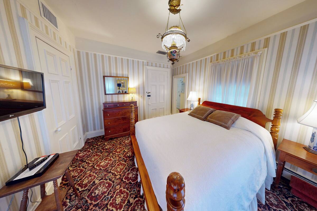 A hotel room with a large bed, a TV, a wooden dresser, a carpeted floor, and a chandelier.