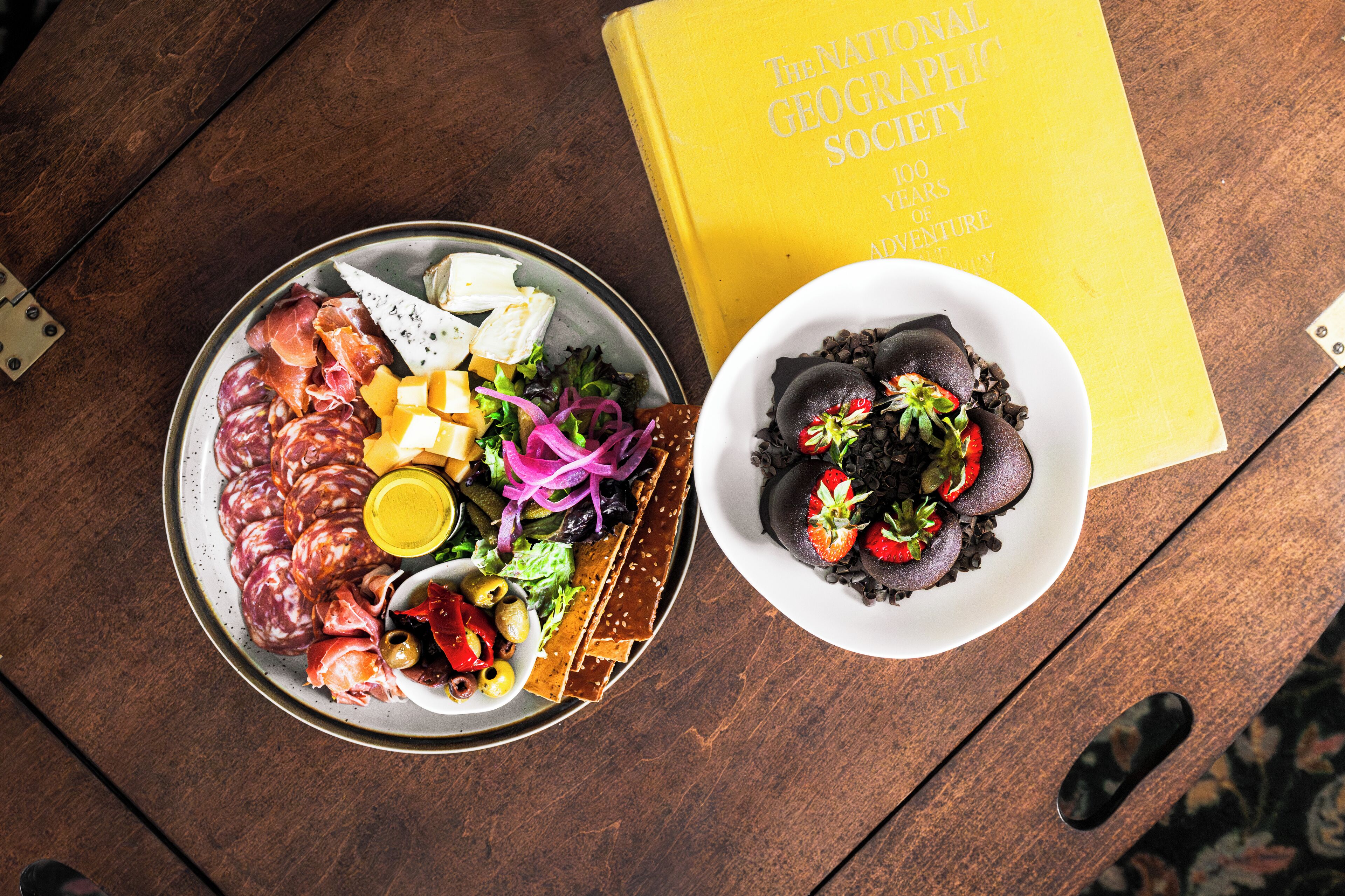 A table with a plate of assorted cheeses, meats, and vegetables, and a bowl of chocolate-covered strawberries. A book titled 'The National Geographic Society 100 Years of Adventure' is also visible.