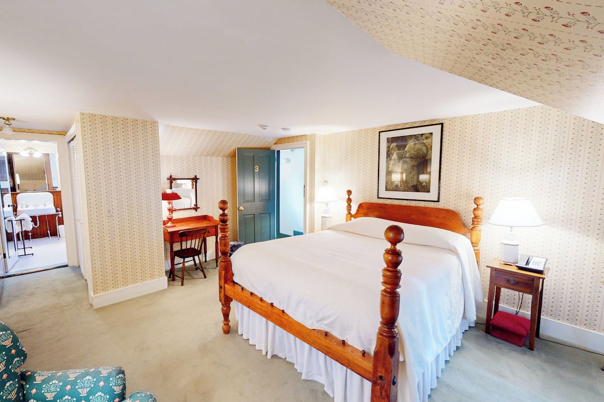 A hotel room with a large bed, a desk, a chair, a small table, a nightstand, a mirror, and a door.