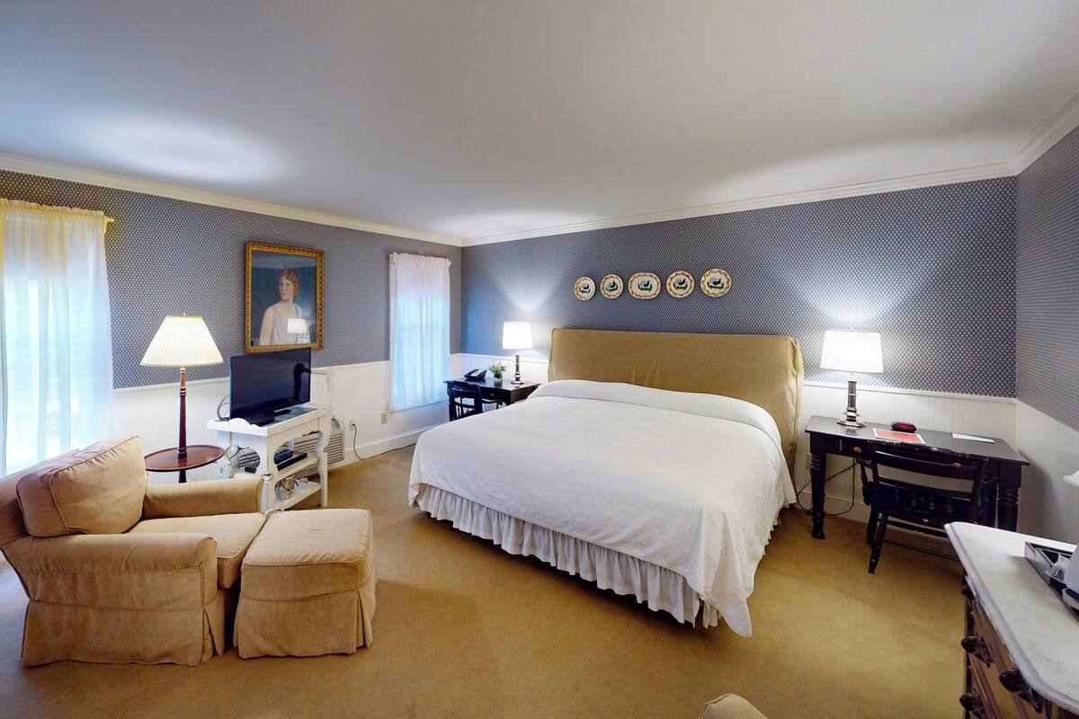 A hotel room with a large bed, a desk, a chair, a TV, and a lamp.