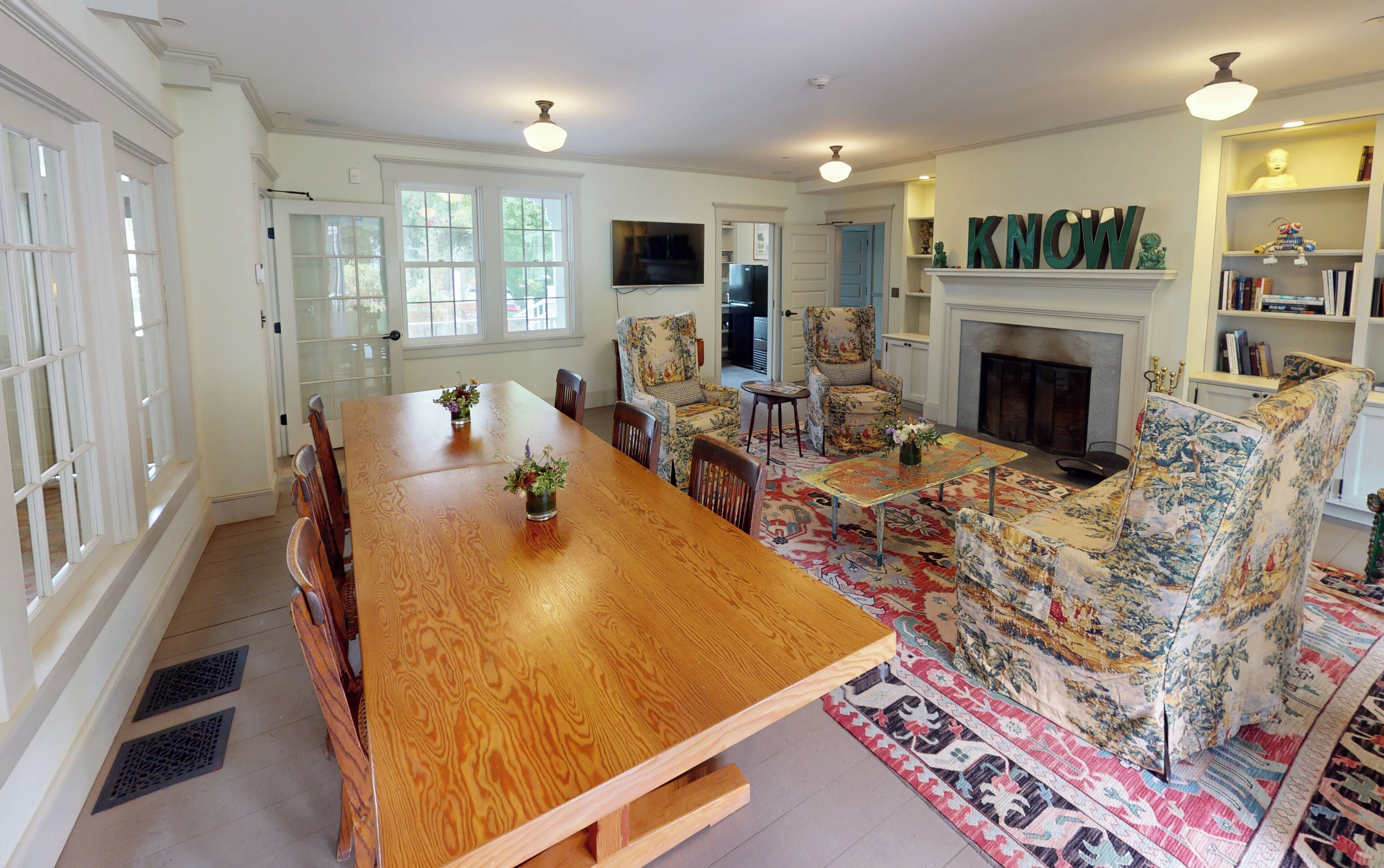 A spacious dining area with a wooden table and floral-patterned chairs, a fireplace, and a bookshelf with decorative items.