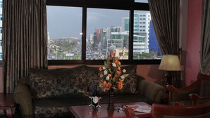 A hotel room with a sofa, a table with a vase of flowers, a lamp, and a view of a cityscape through large windows. - Haimi Apartment Hotel (Addis Ababa)