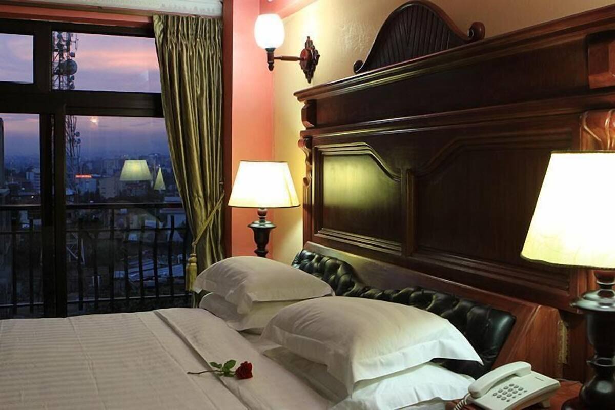 A hotel room with a large bed, two bedside lamps, a telephone, and a view of the city through a large window.