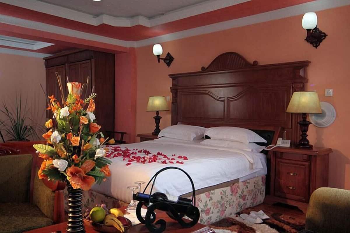 A hotel room with a large bed, two bedside lamps, a wooden headboard, a nightstand with a lamp, a vase with flowers, and a small table with fruits and a book.