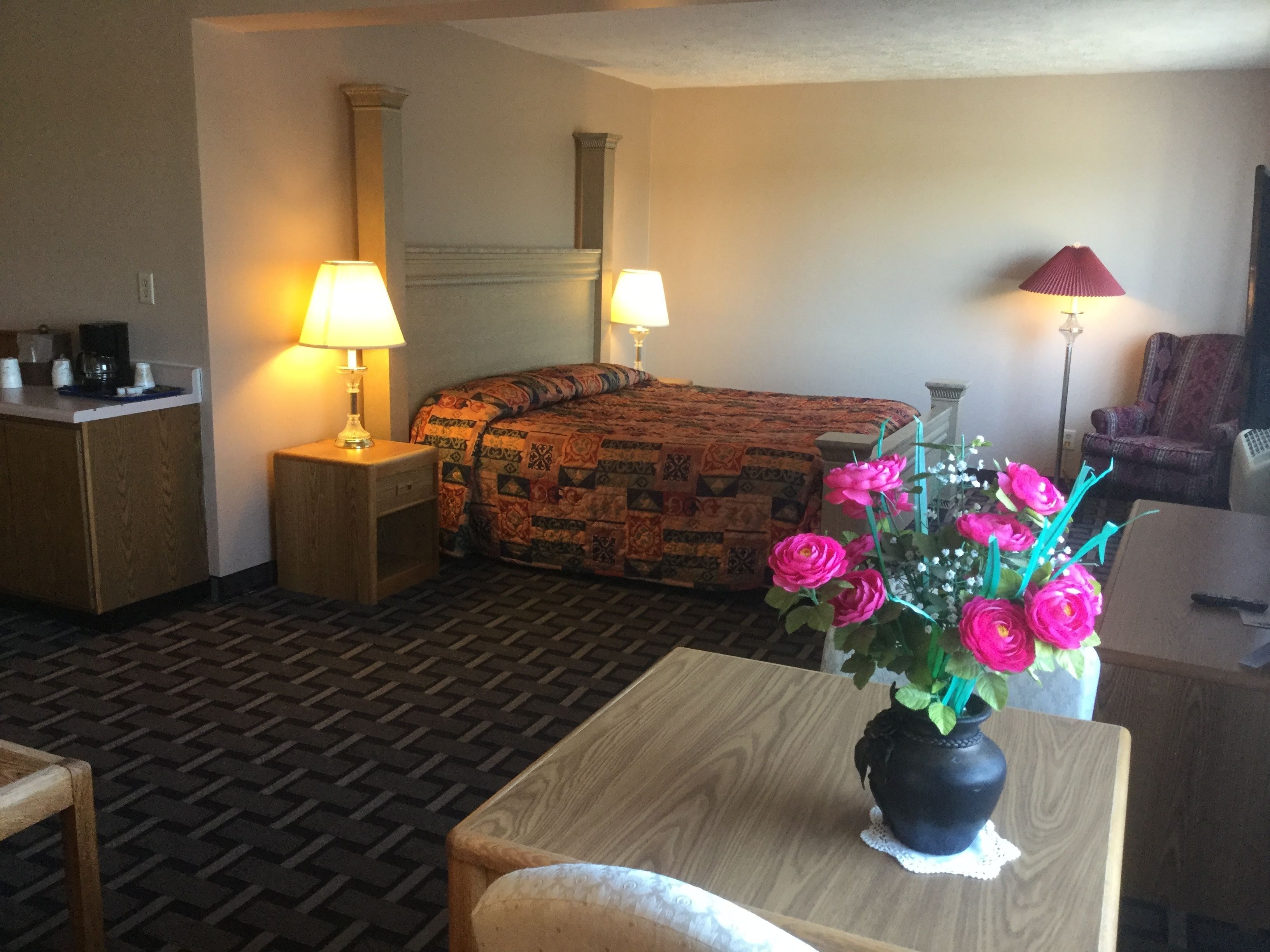 Una habitación de hotel con una cama, dos lámparas de noche, una mesita con un florero, una silla, un escritorio y una cafetera.