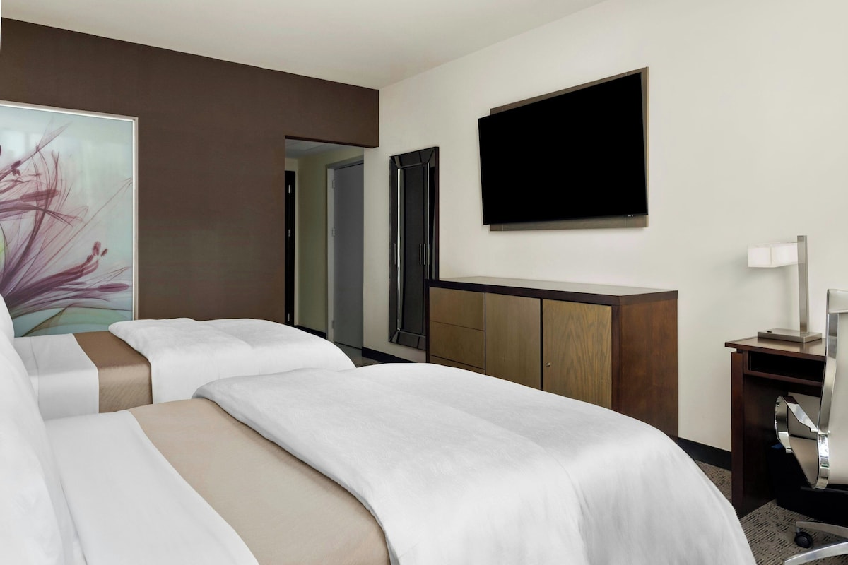 Habitación de hotel con dos camas, un televisor de pantalla plana, un escritorio con lámpara y un cuadro en la pared.