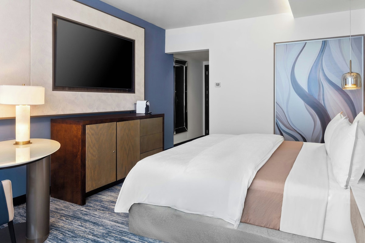 Una habitación de hotel con cama, escritorio con lámpara, televisión y un cuadro en la pared.