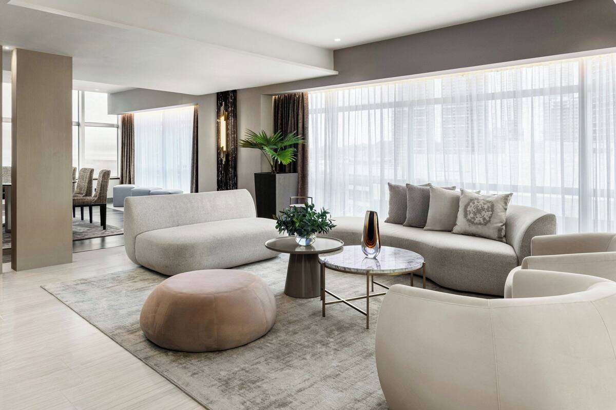 Un salon moderne avec un canapé, un pouf et une table basse, de grandes fenêtres avec des rideaux légers et une vue sur la ville.