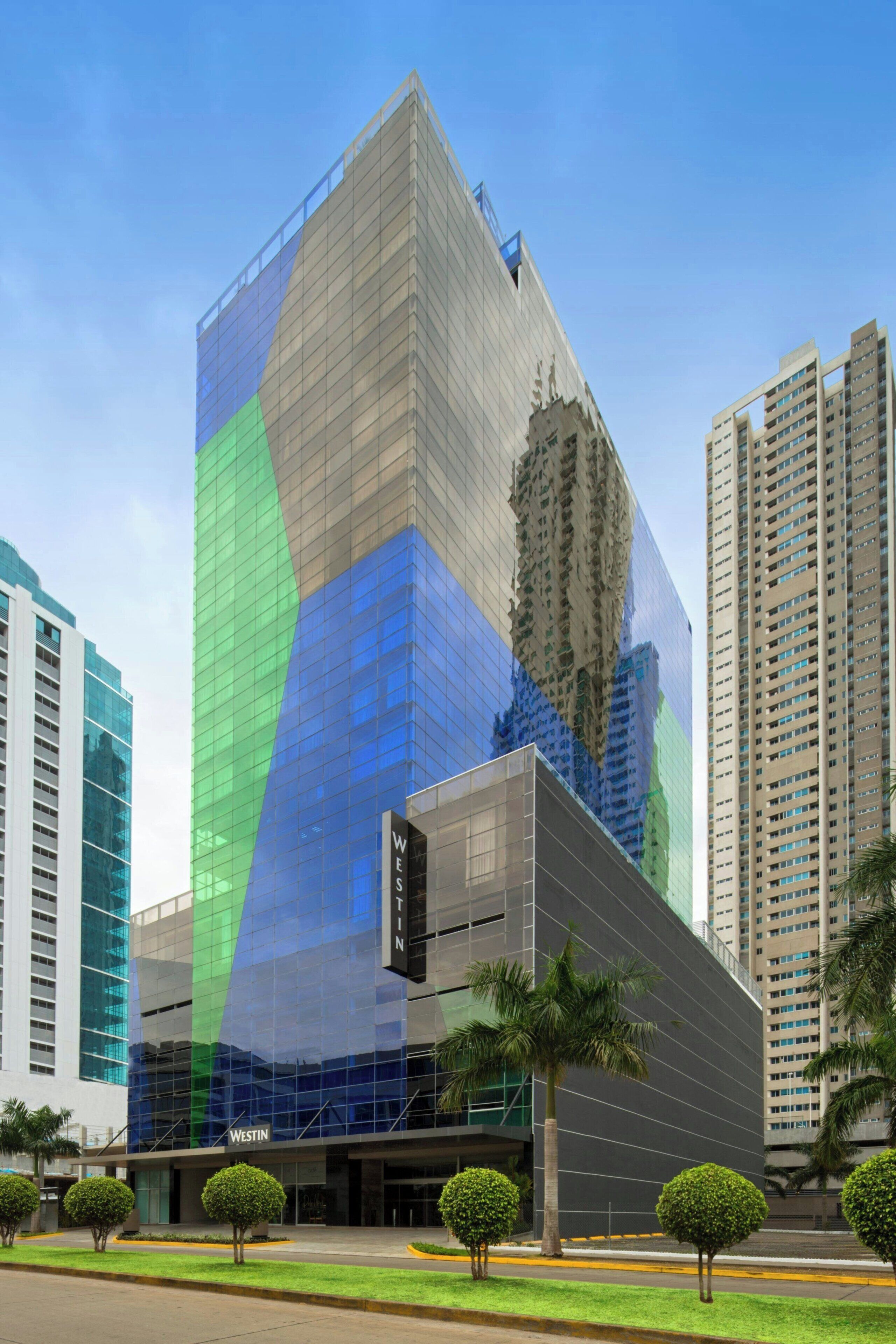 A modern hotel with a distinctive geometric facade, surrounded by high-rise buildings and palm trees.