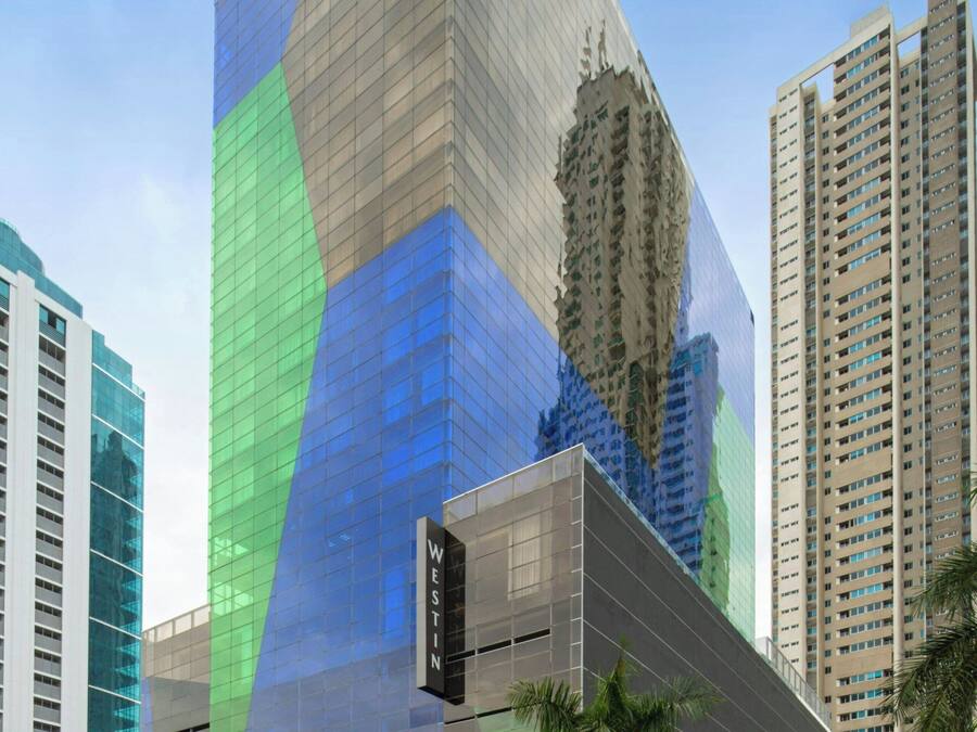 A modern hotel with a distinctive geometric facade, surrounded by high-rise buildings and palm trees.