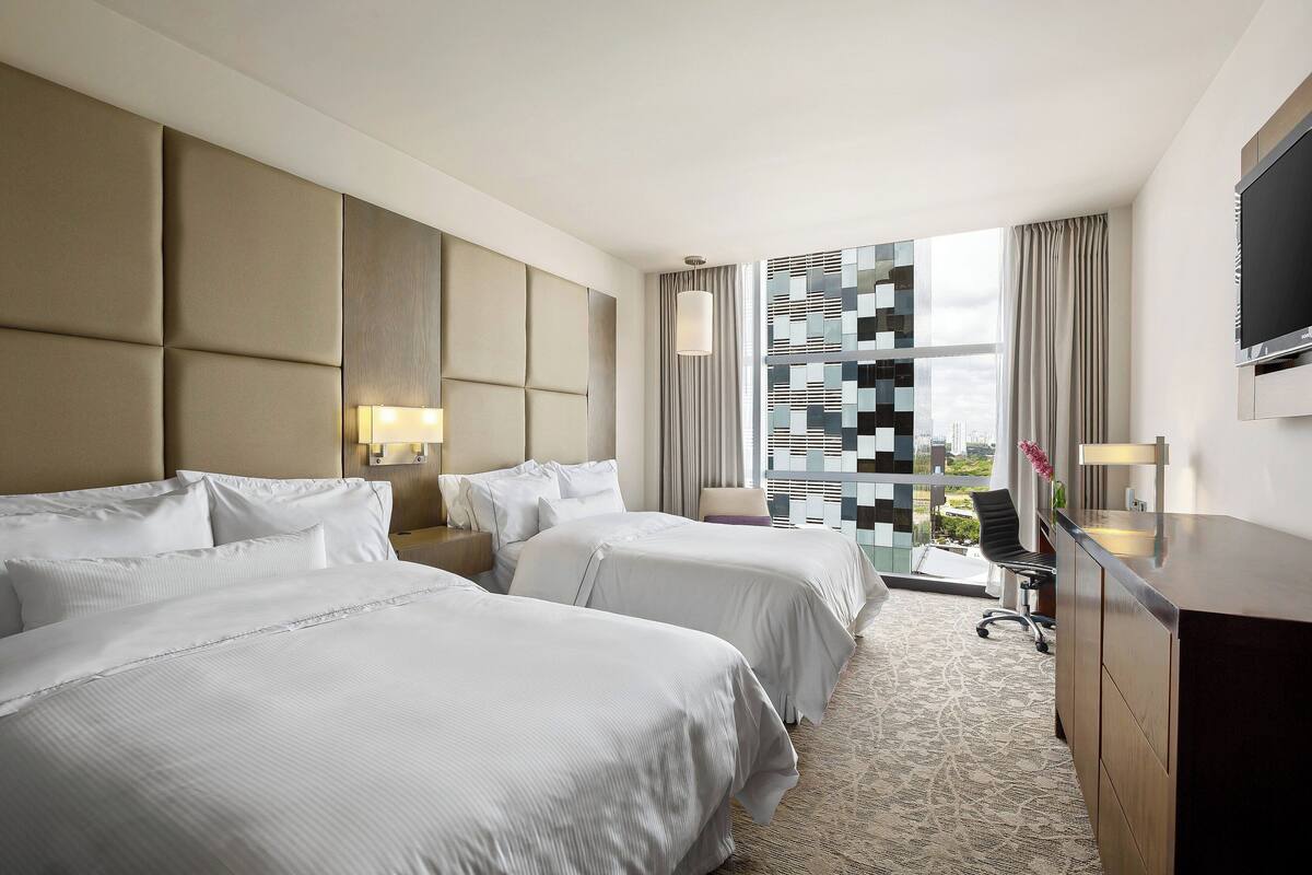 Une chambre d’hôtel avec deux lits, un bureau, une télévision et une vue sur la ville grâce à une grande fenêtre.