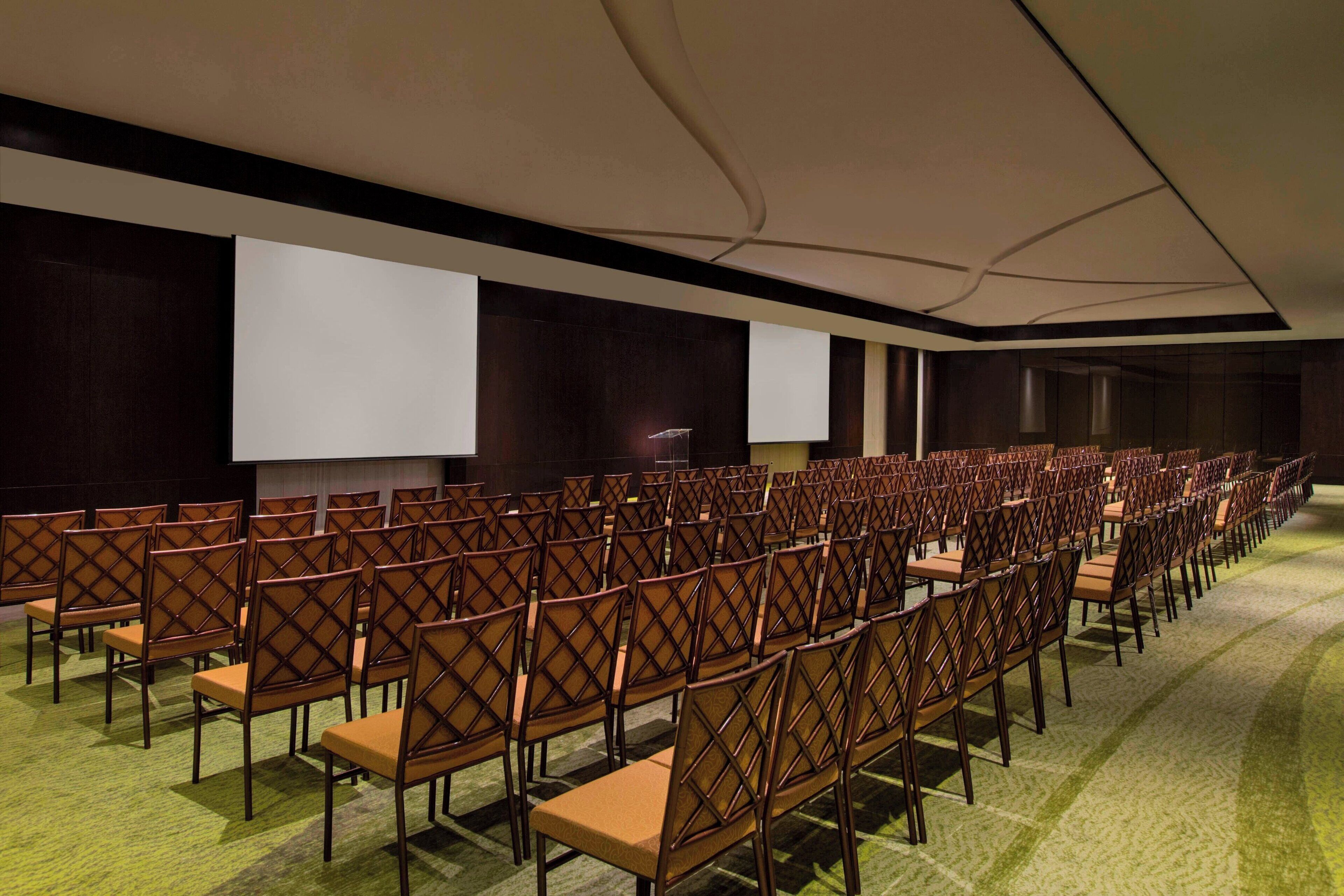 A conference room with rows of wooden chairs, a large screen, and a carpeted floor.