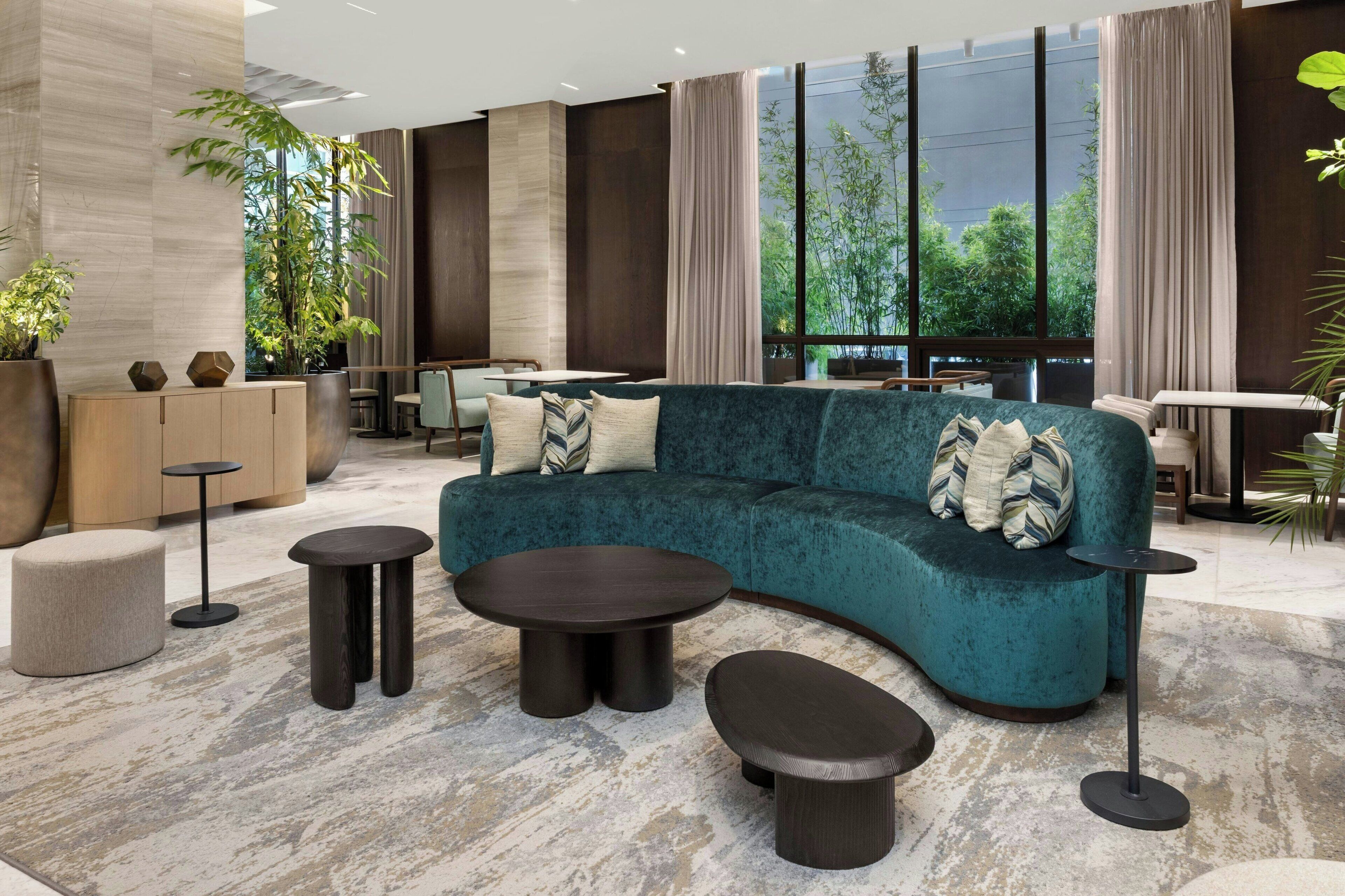 Et moderne opholdsområde med en teal sofa, runde sofaborde og et stort vindue med udsigt til grønne områder.