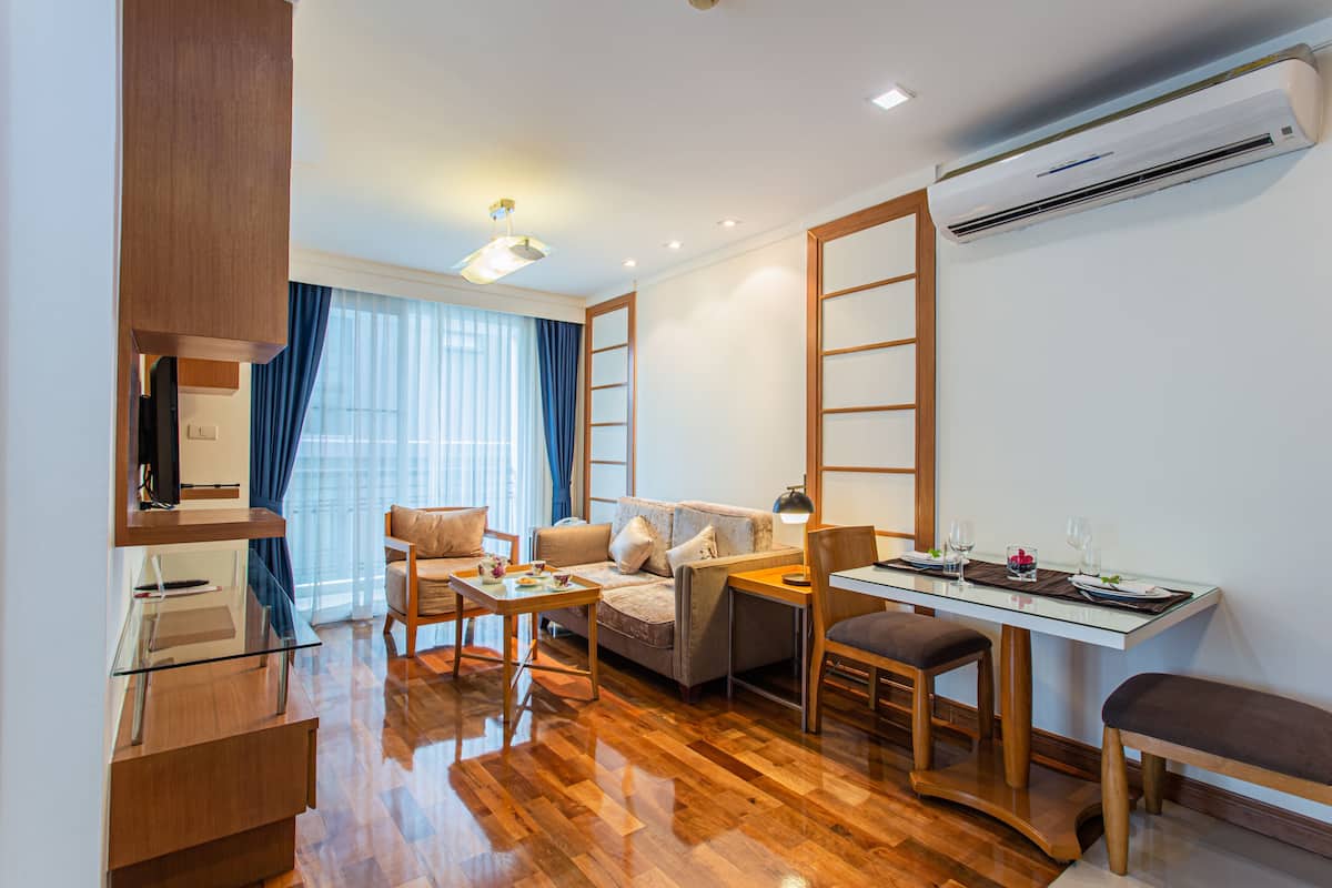 A modern hotel room with a wooden floor, a sofa, a dining table with chairs, a TV, and an air conditioning unit.