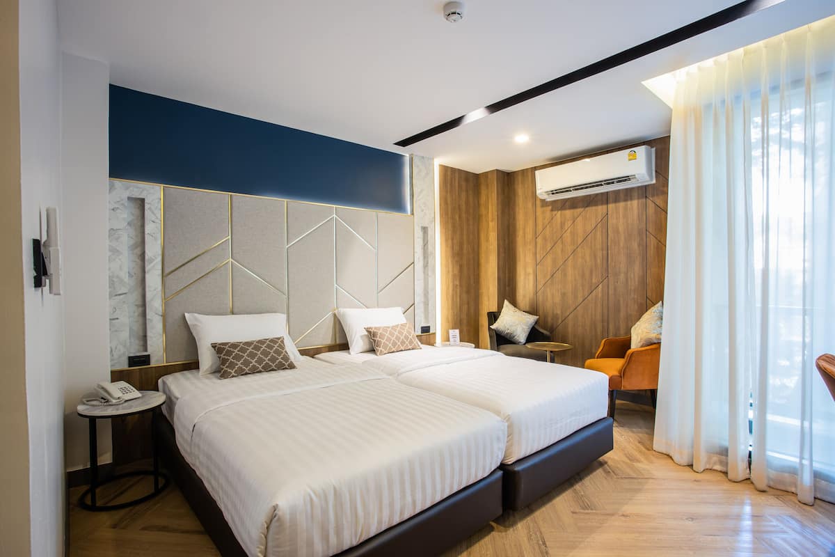 A modern hotel room with two beds, a wooden accent wall, an air conditioning unit, and a seating area with a chair and table.
