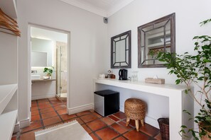 A modern bathroom with a white vanity, two mirrors, a small black trash bin, a wicker stool, and a potted plant.
