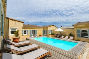 Une piscine dotée d’une terrasse en bois et de chaises longues, entourée de bâtiments aux volets bleus.