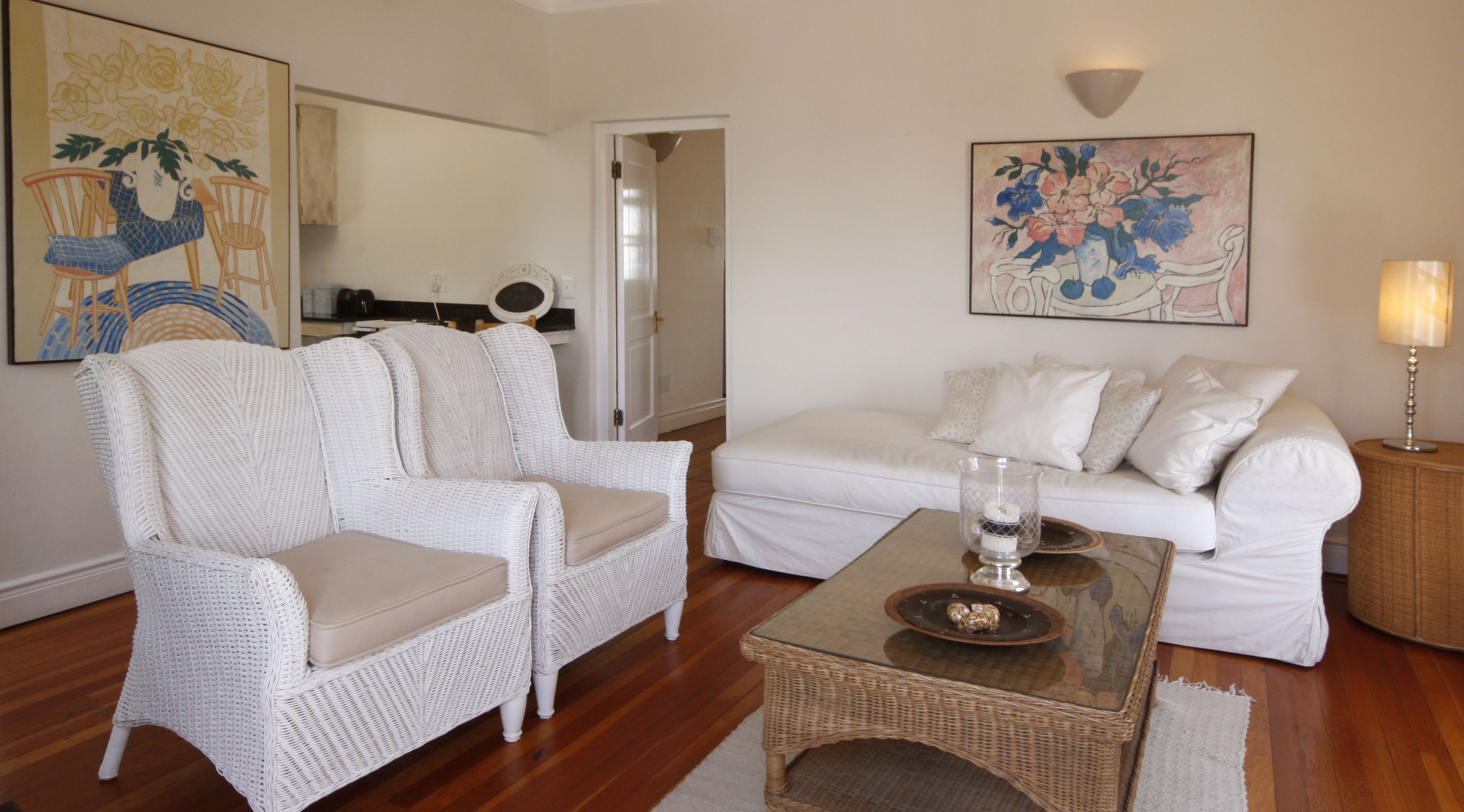 Un salon avec un canapé blanc, deux fauteuils en osier blanc, une table basse en verre et une table d’appoint en osier. Deux tableaux sont accrochés aux murs, une porte se trouve au fond et une lampe est posée sur la table d’appoint.