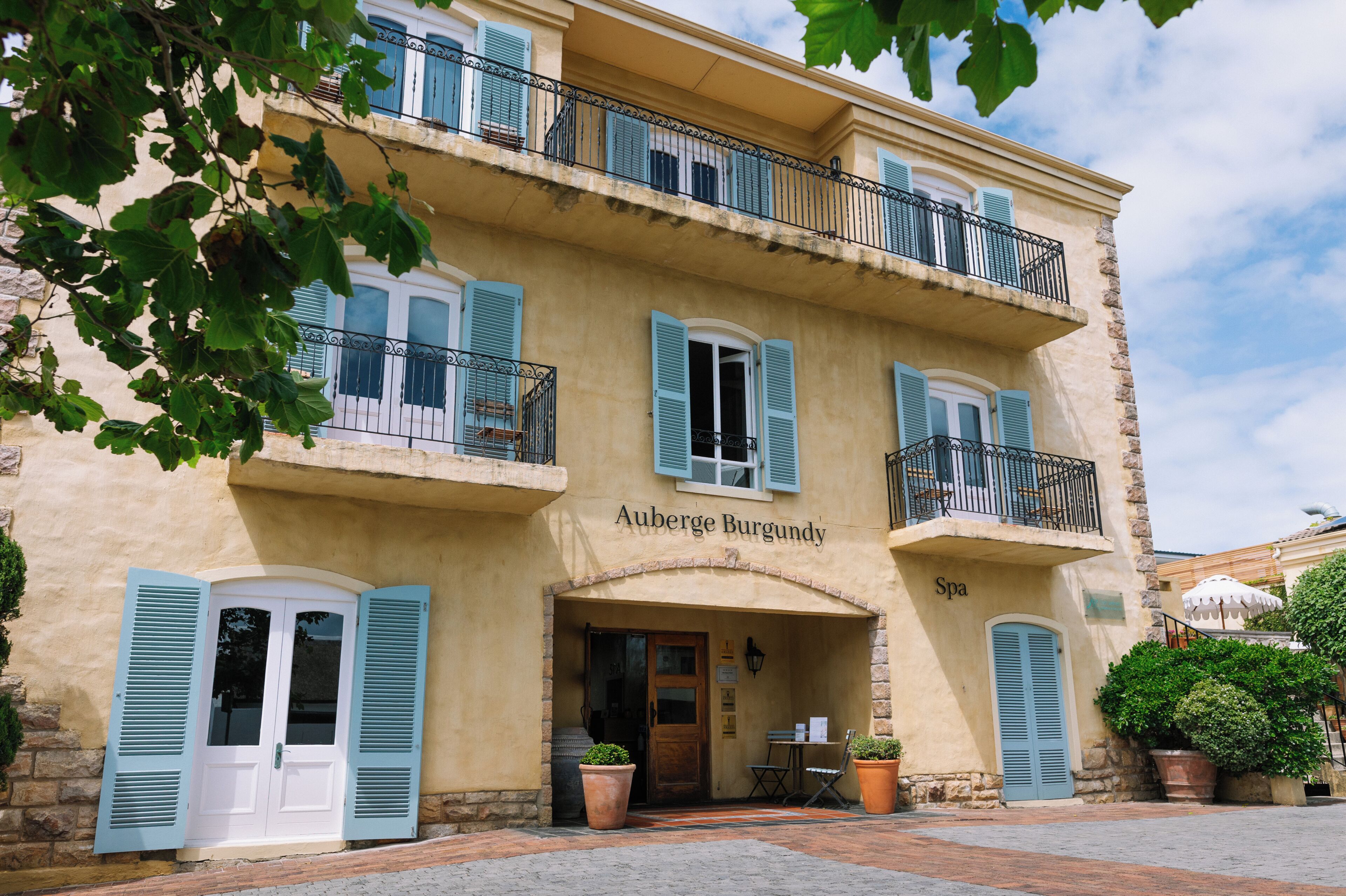A building with a sign reading 'Auberge Burgundy Spa,' featuring a stone facade, blue shutters, and a balcony with a wrought iron railing.