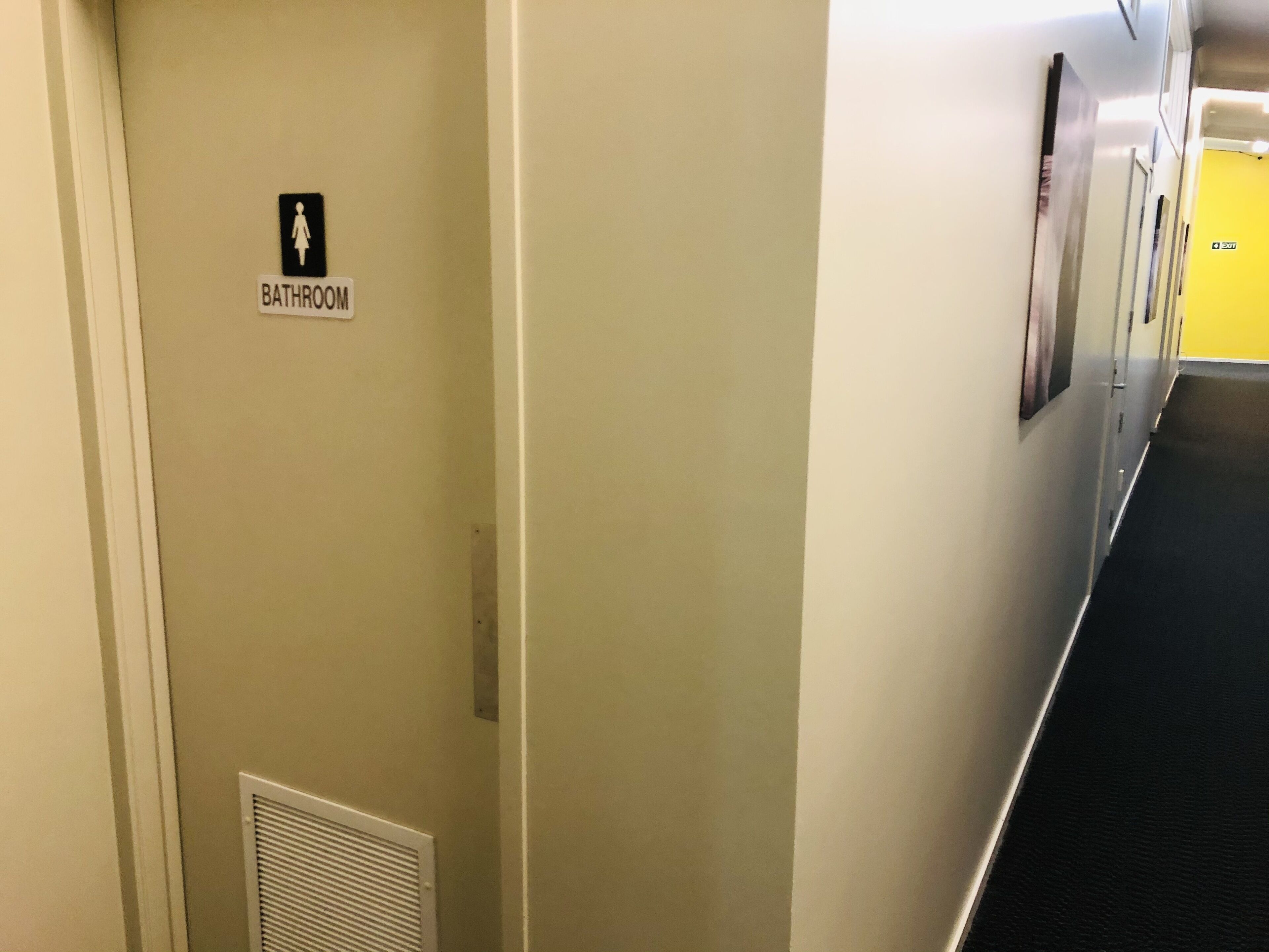 Pasillo de hotel con un letrero que indica un baño, una rejilla de ventilación y una puerta amarilla al final.