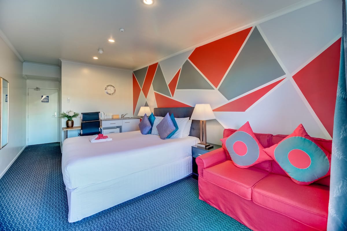 A hotel room with a large bed, a red sofa, a desk with a computer, and a wall with geometric patterns.