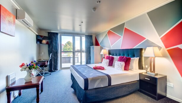 A modern hotel room with a large bed, a desk, a chair, a TV, and a view of the outdoors. - Silver Fern Lodge (Taupo)
