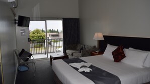 A hotel room with a bed, a desk, a chair, a TV, and a balcony with a view of trees and buildings. - Silver Fern Lodge (Taupo)
